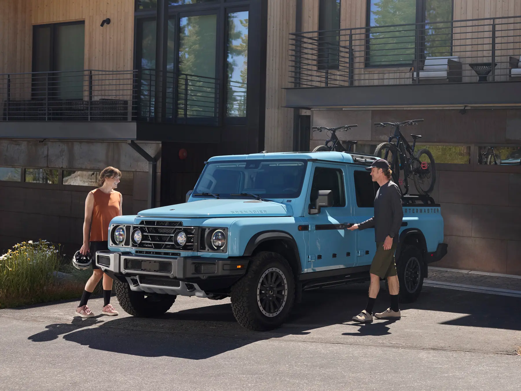 Light blue INEOS Grenadier Quartermaster pickup on a driveway with mountain bikes carried on a bed-mounted rack, two people walking towards it
