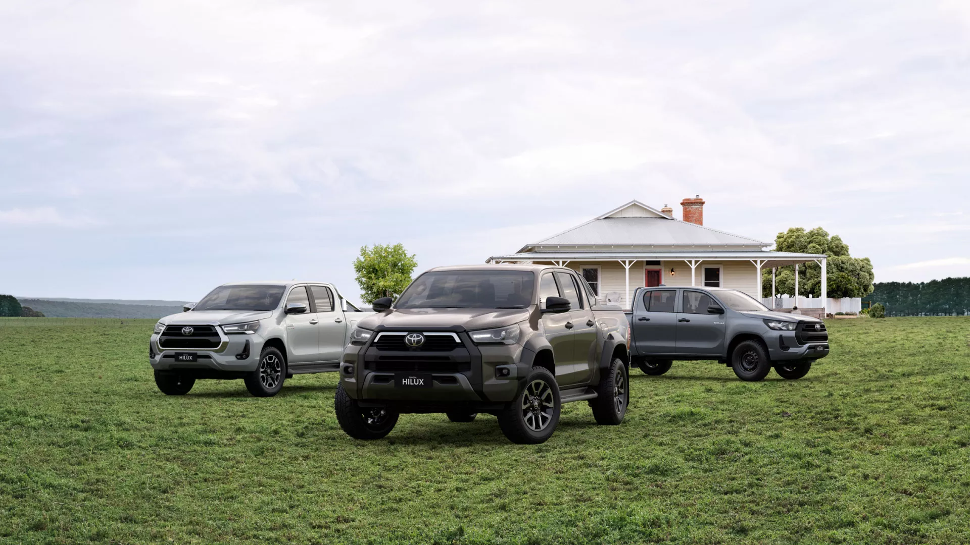 Three Toyota Hilux dual-cabs in silver, bronze and grey parked on grass in front of a weatherboard farmhouse