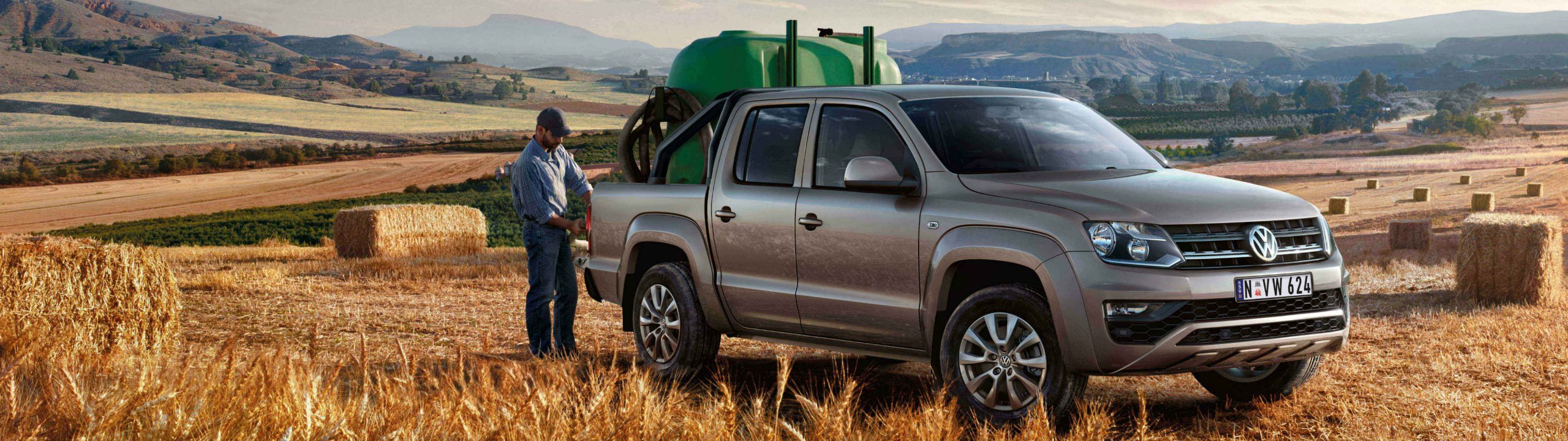Volkswagen Amarok dual-cab carrying a green water tank in its tub, parked in a harvested field with a man at the tailgate